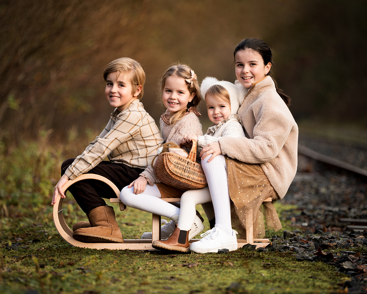 Fotograf Bergen. Familiefotograf. Søskenbilder. 4 søsken. beige klær. fire barn sitter på en kjelke. Høstfarger ute i bakgrunnen.