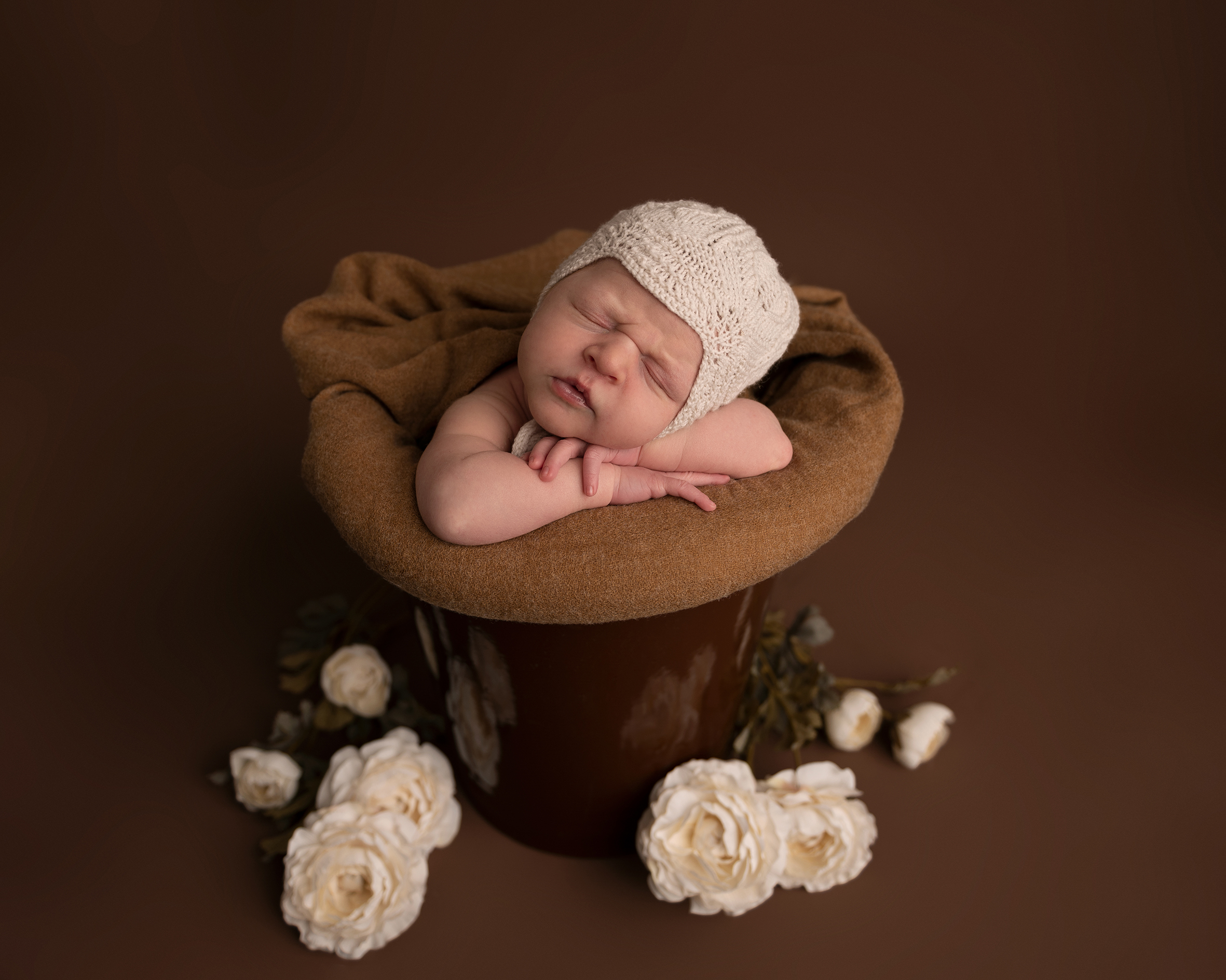 Nyfødtfotograf i Bergen. Bilder av baby. Nyfødtfotografering. Nyfødtbilder. Fotograf Maria. Gutt med hvit strikkelue i en brun bøtte.Brun bakgrunn med hvite ranukler blomster.